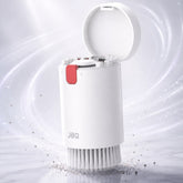 A white cylindrical cleaning brush with an open lid, shown on a light marble-like surface with a soft white and gray background and swirling light effects. The brush has a small red tab and the brand name JBQ printed on the front.