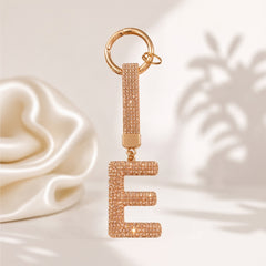 A rose gold letter 'E' keychain charm paved with small clear rhinestones, featuring a matching rhinestone-embellished strap and a gold-tone split ring, displayed against a soft white background with decorative shadows.