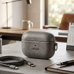 A textured gray protective case for AirPods sits on a gray stone desk in a modern, softly lit home office. The case features the 'piblue' brand logo.