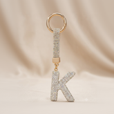 A gold-tone keychain featuring a rhinestone-encrusted letter 'K' charm and a matching rhinestone strap, set against a soft beige fabric background.