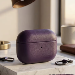 A purple leather AirPods case sits on a white marble block on a desk, surrounded by a brass desk lamp, a coffee mug, a notebook, and a matching purple leather charging cable with a USB-C connector.