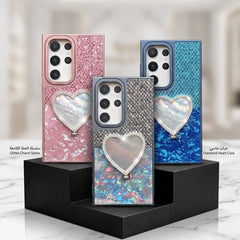 Three smartphone cases displayed on black stands in a clean indoor setting. The center case is shown in front, with a glittery iridescent heart charm and a blue patterned back.