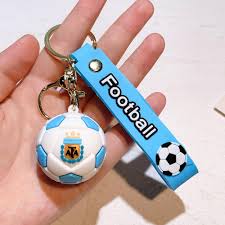 A hand holding a small soccer-themed keychain with a white and light blue soccer ball charm, a blue strap labeled 'Football', and a metal key ring.