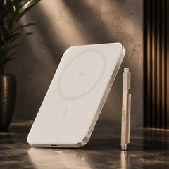 A white wireless charging pad with a circular charging symbol on its surface, shown standing upright on a dark marble surface next to a metallic pen. The background features a textured gray wall with warm sunlight shadows and a potted plant on the left.