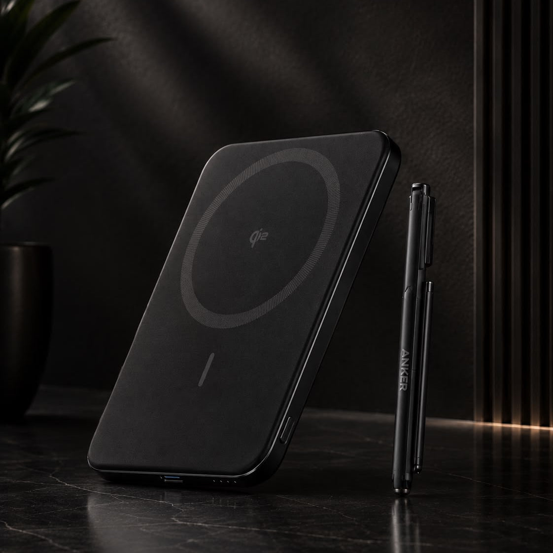 A black magnetic wireless charging pad with a circular charging symbol, shown standing on a dark marble surface next to a slim black stylus pen in a moody, dark interior setting.