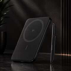 A black magnetic wireless charging pad with a circular charging symbol, shown standing on a dark marble surface next to a slim black stylus pen in a moody, dark interior setting.