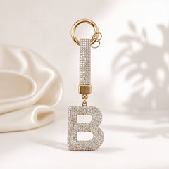 A gold-tone initial letter 'B' keychain charm covered in clear rhinestones, with a matching rhinestone-embellished strap and split key ring, shown on a soft cream background.