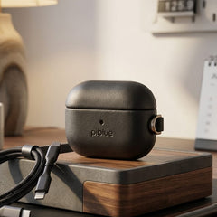A dark gray piblue wireless earbud charging case sits on a wooden and gray stand on a desk, with a braided charging cable nearby in a warm, modern home office setting.