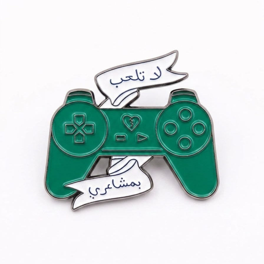 A green video game controller enamel pin with silver metal outlines and two white banners with Arabic text, shown on a plain white background.