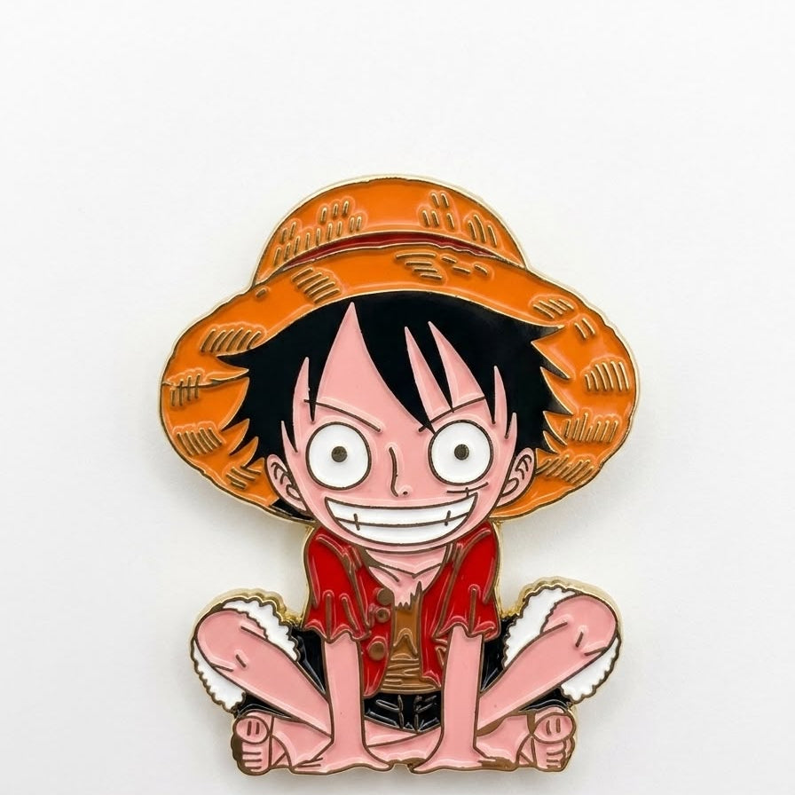 An enamel pin of the anime character Monkey D. Luffy from One Piece, sitting cross-legged and wearing his signature orange straw hat and red shirt, set against a plain white background.
