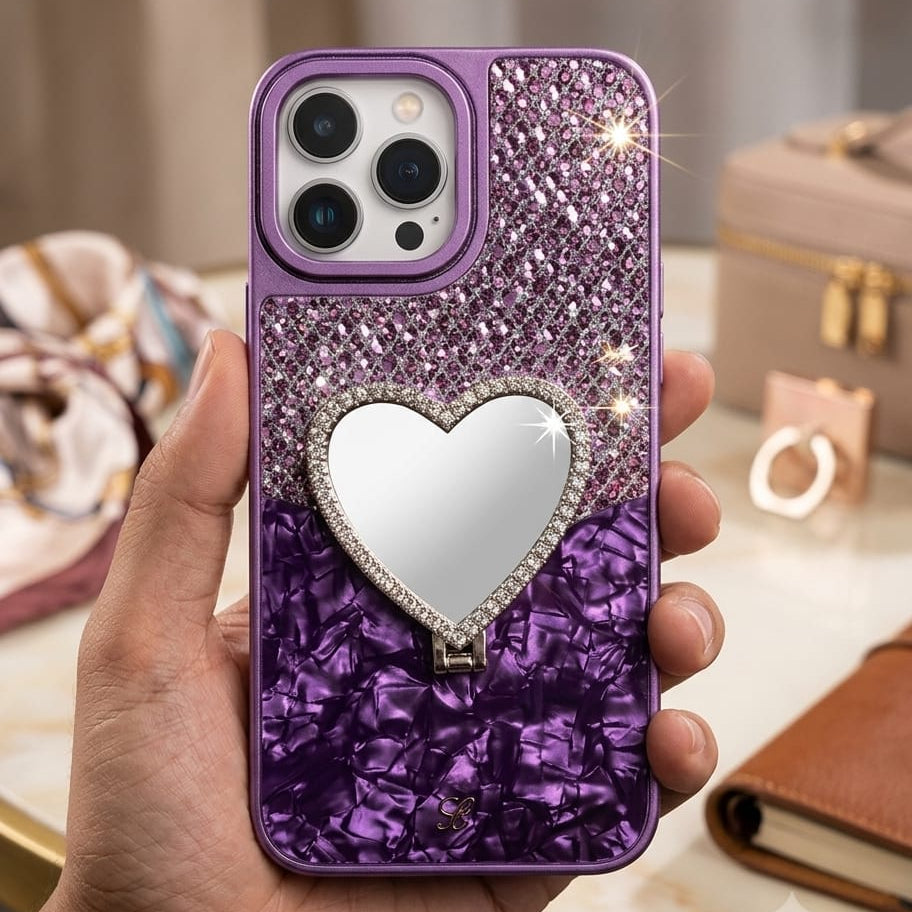 A hand holds a purple glitter phone case decorated with a heart-shaped mirror and rhinestone trim, set against a softly blurred indoor background.