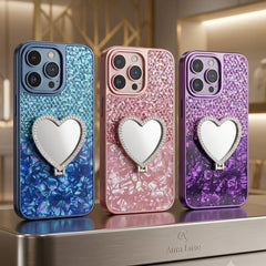 Three glittery phone cases with heart-shaped mirror covers are displayed on a metallic stand. The cases feature blue, pink, and purple gradient glitter finishes with a central silver heart mirror.