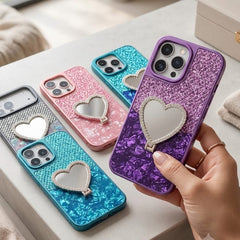 A hand holds a purple glitter phone case with a heart-shaped mirror, while four more matching phone cases in silver, gray, blue, pink, and purple are arranged on a tabletop in a bright indoor setting.