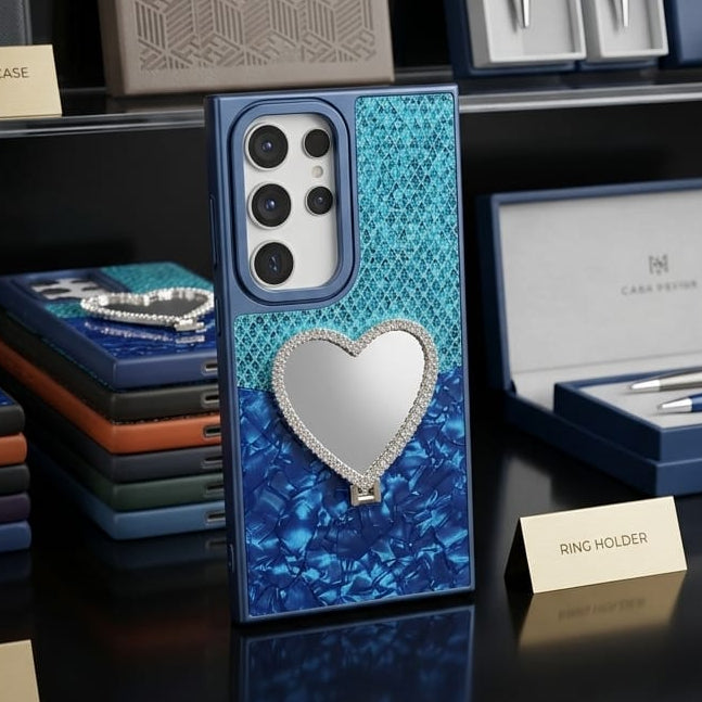 A blue textured phone case with a large rhinestone-embellished heart on the back, displayed on a black shelf alongside branded packaging and accessories.