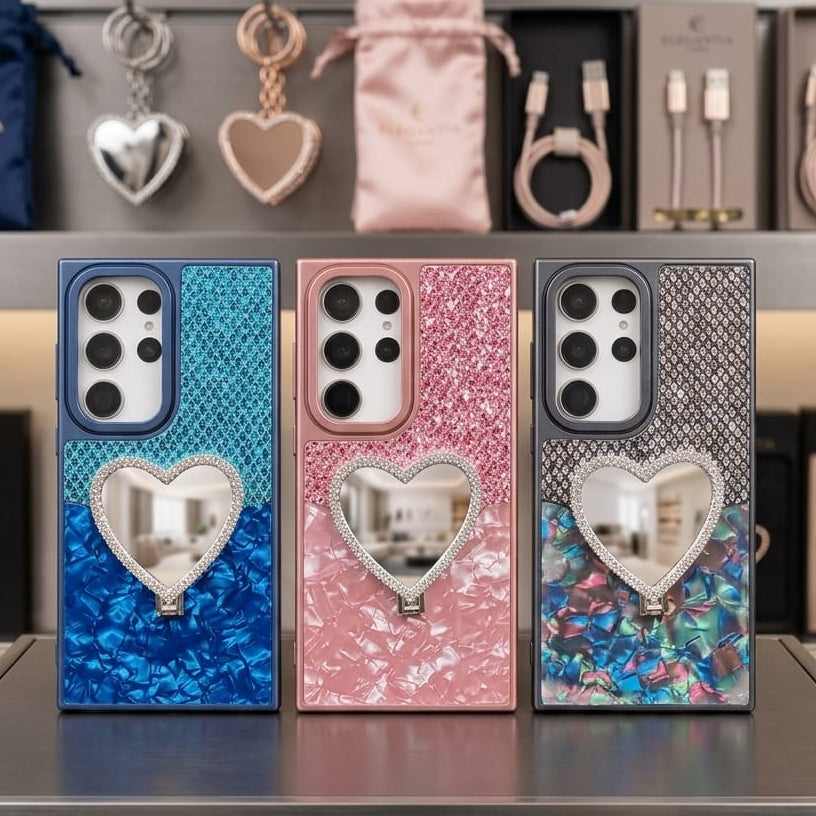 Three elegant phone cases with heart-shaped mirror inlays are displayed on a counter. The left case is blue and silver textured, the middle is pink and silver glitter, and the right is dark gray and multicolor marbled with a silver heart.