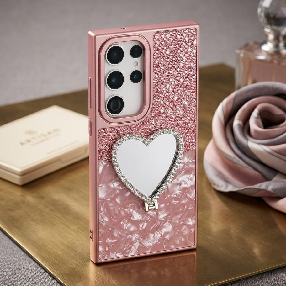 A pink glitter and pearl-pattern phone case with a heart-shaped mirror and rhinestone trim, shown standing on a gold tray with decorative props in a styled setting.